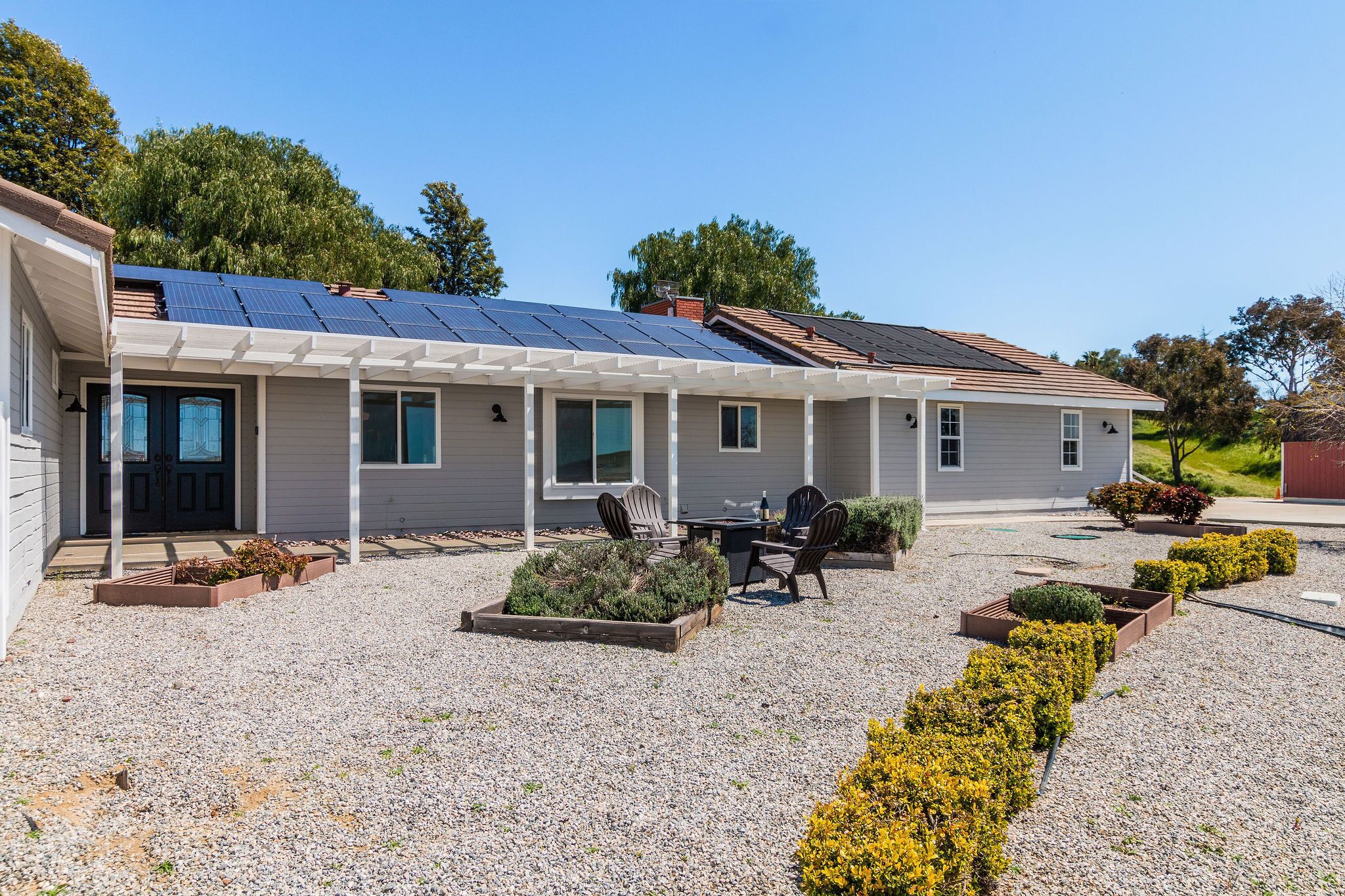 A single-story house with solar panels on the roof, a covered patio with outdoor furniture, and a gravel driveway with landscaped garden beds.