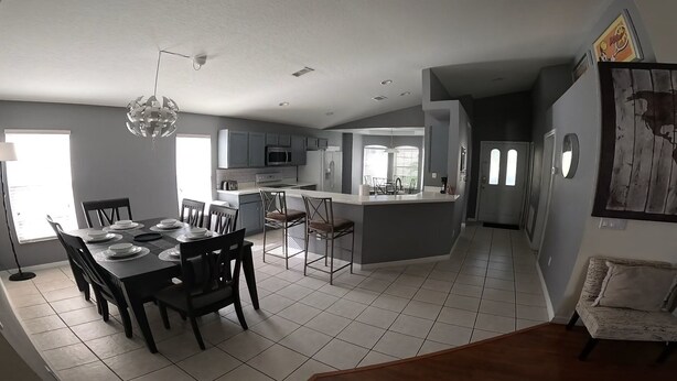 A modern kitchen with grey cabinets, a central island, and a dining area with a table set for four. - WOW! Championsgate-Minutes to disney,private pool,wifi ,tennis,gym,games room  (Davenport)