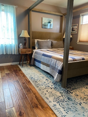 A four-poster bed with a quilt, a nightstand with a lamp, a patterned rug, and wooden flooring.