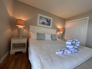 Un dormitorio ordenado con una cama grande, dos mesitas de noche con lámparas, un cuadro en la pared y una puerta al fondo.