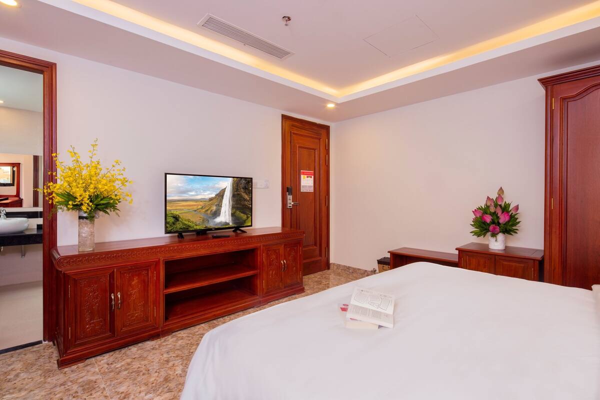 Quarto de hotel com uma cama grande, televisão sobre suporte de madeira, guarda-roupa de madeira e uma pequena mesa com um vaso de flores.