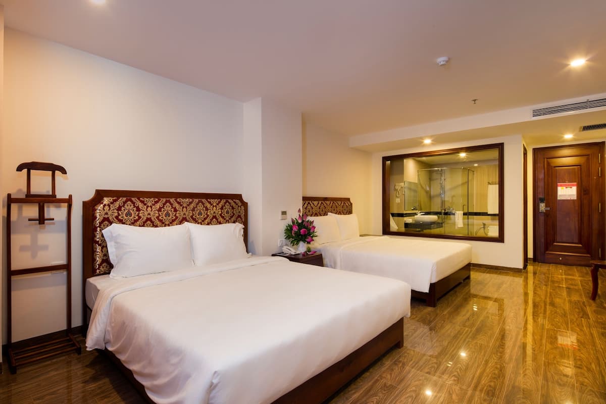 Quarto de hotel com duas camas, cabeceira de madeira, mesa de cabeceira com abajur e um espelho grande.