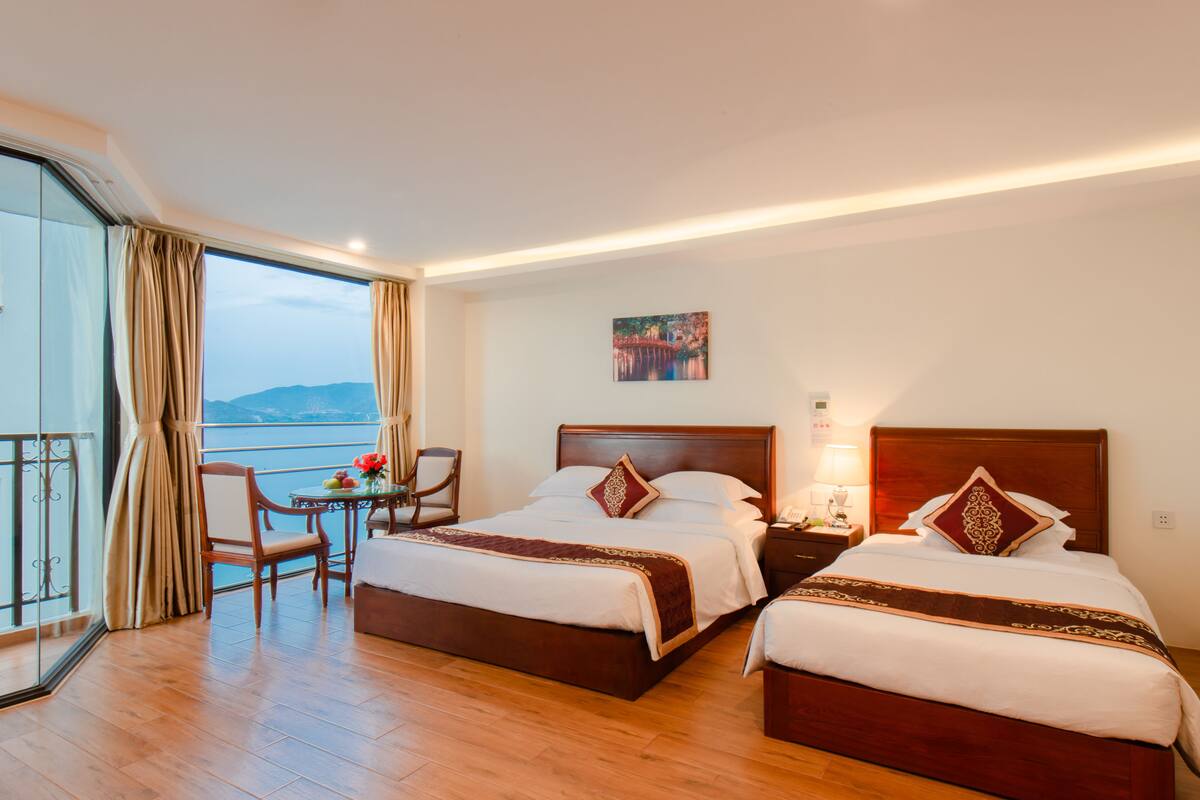 Quarto de hotel com duas camas, varanda com vista, uma mesa pequena com cadeiras e um quadro na parede.