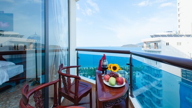 A balcony with a table set with fruit and a glass of wine, overlooking a cityscape and the sea. - Red Sun Nha Trang Hotel (Nha Trang)