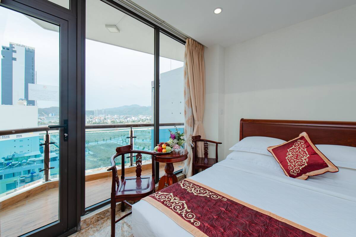 Quarto de hotel com uma cama grande, uma cadeira de madeira, uma mesa pequena com frutas e flores, e uma varanda com vista para a cidade.
