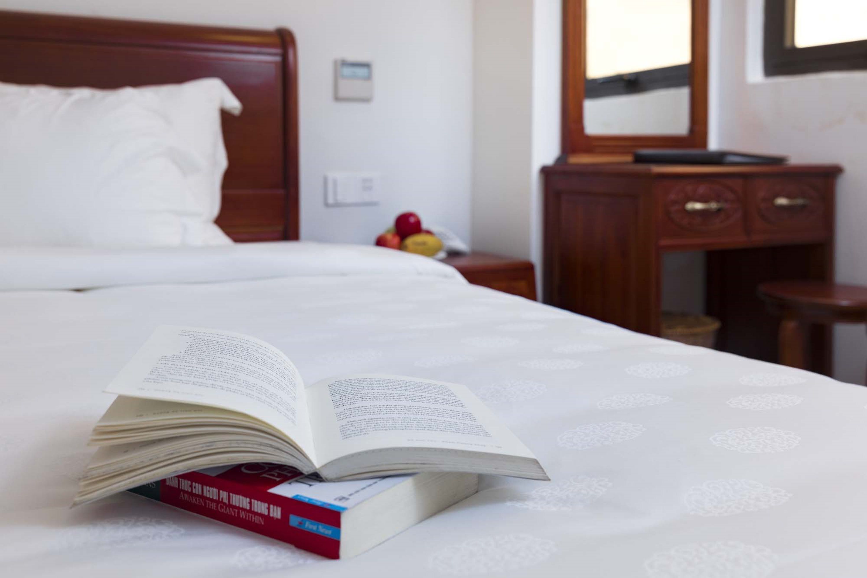 A neatly made bed with a book open on it, a bedside table with a fruit bowl, and a wooden dresser with drawers.