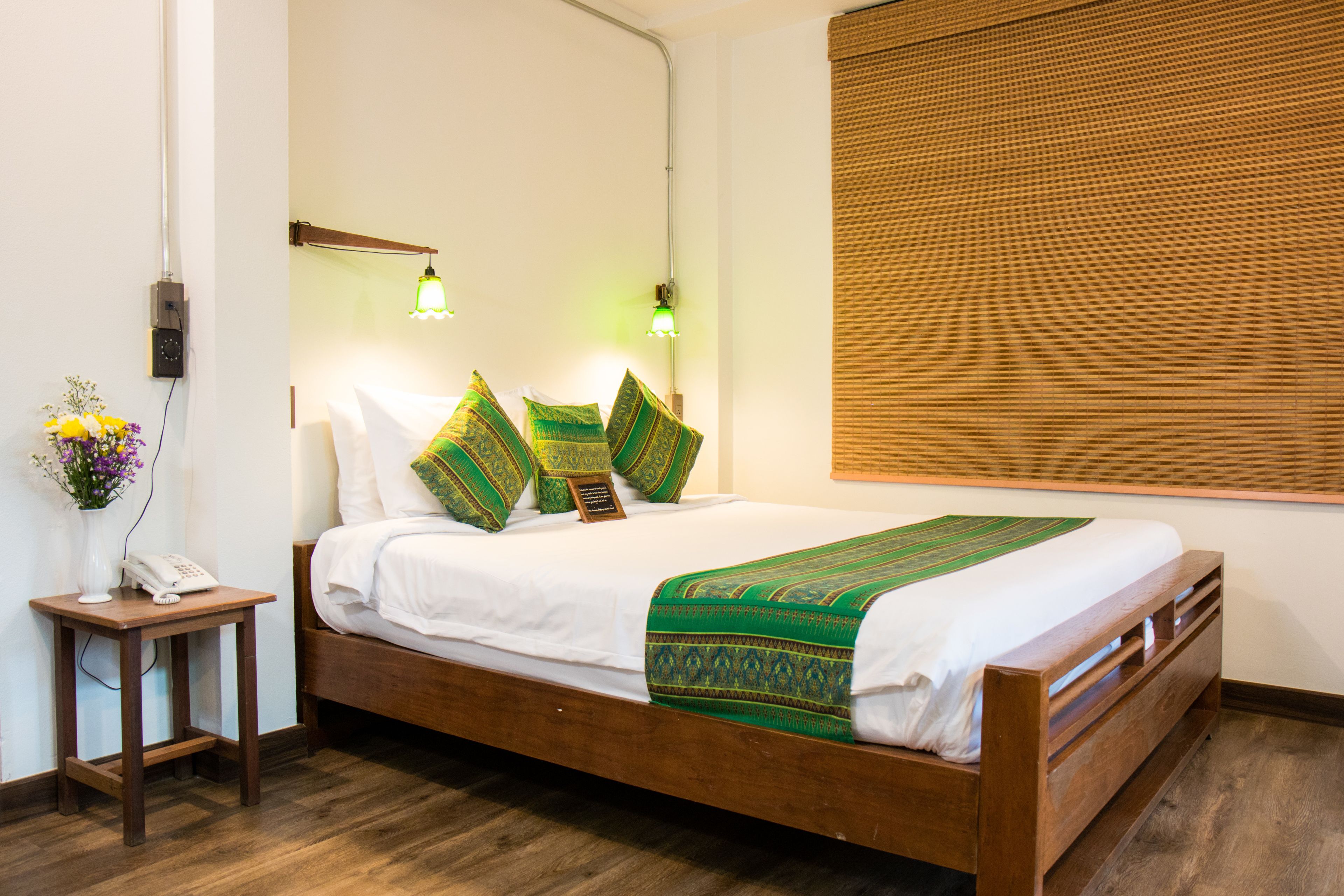 A neatly made bed with a green and white bedspread, a wooden bedside table with a phone, and a wall-mounted lamp.