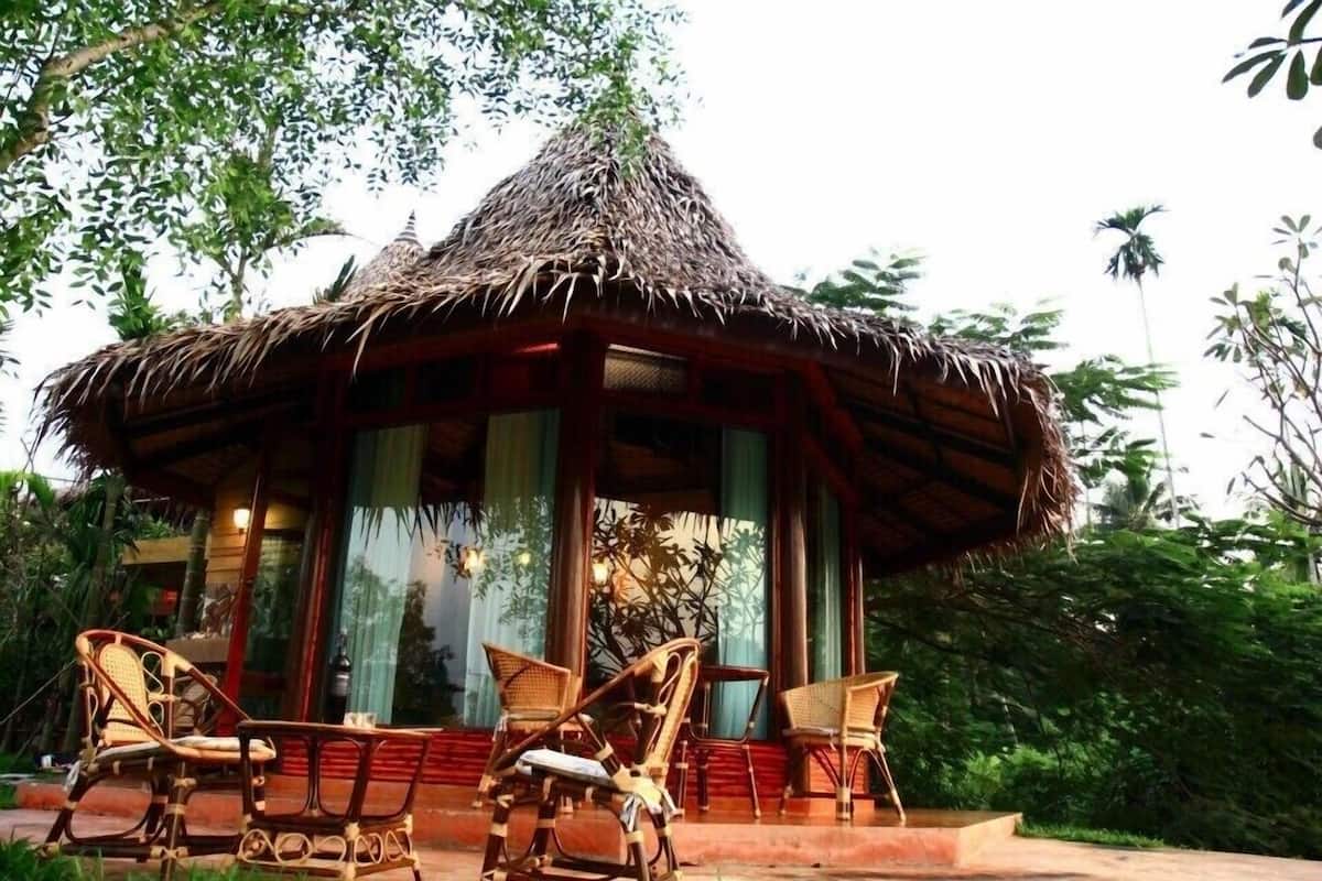 A thatched-roof gazebo with wicker furniture and a glass door, surrounded by lush greenery.