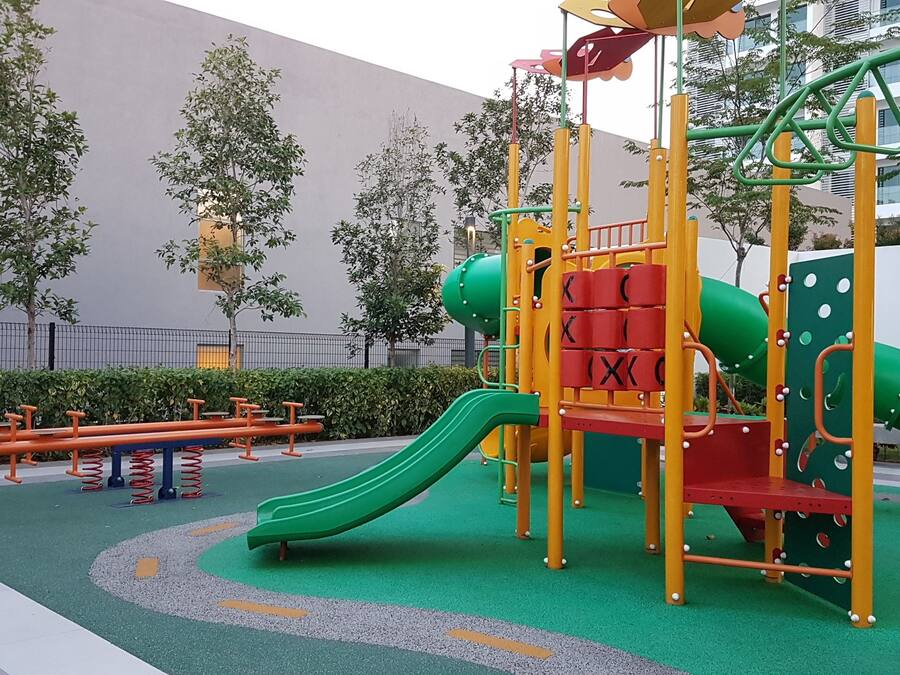 Une aire de jeux avec un toboggan et des structures d’escalade, entourée d’une zone pavée et d’arbres.