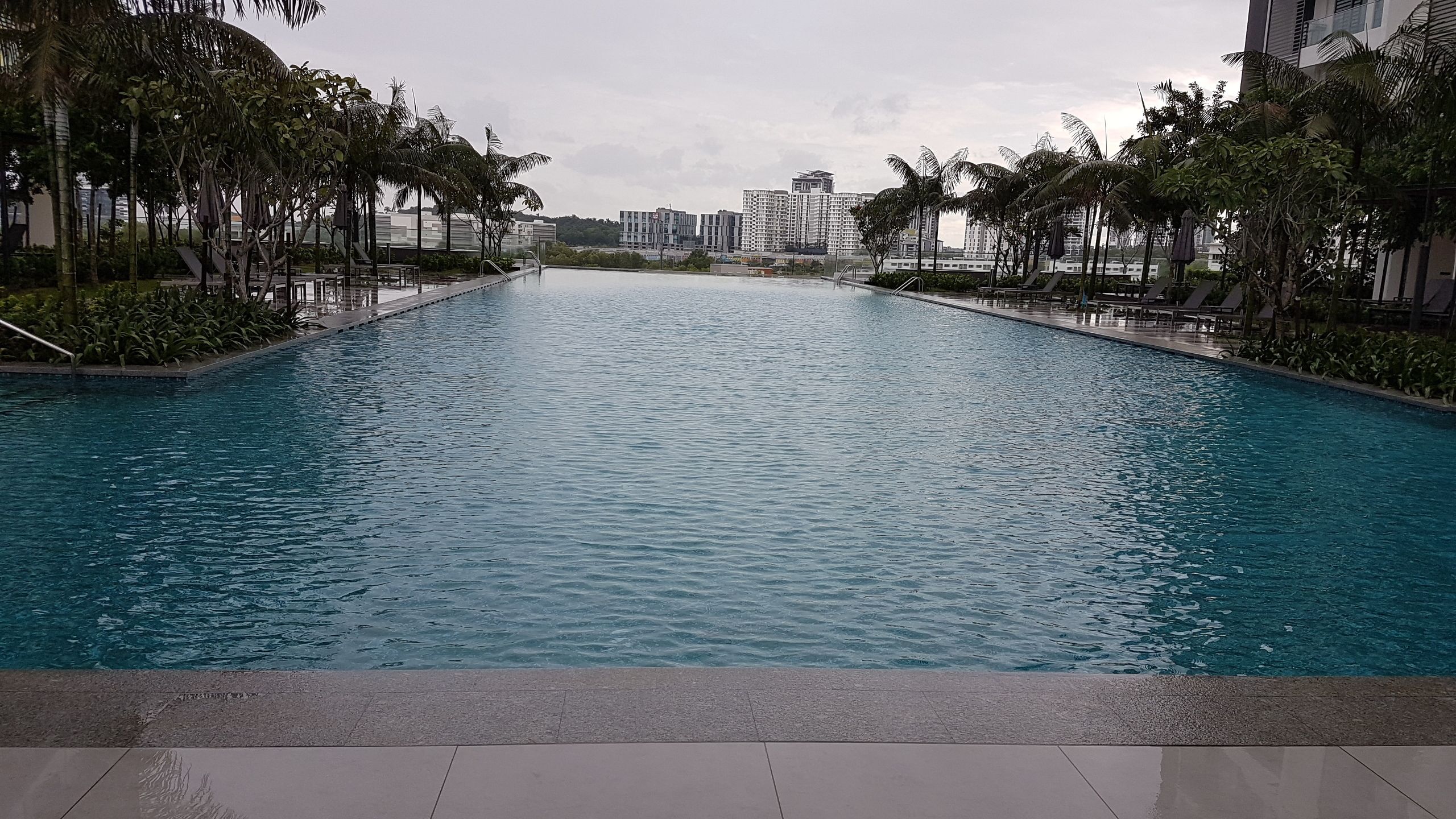 A long, rectangular swimming pool with clear blue water, surrounded by palm trees and modern buildings in the background.