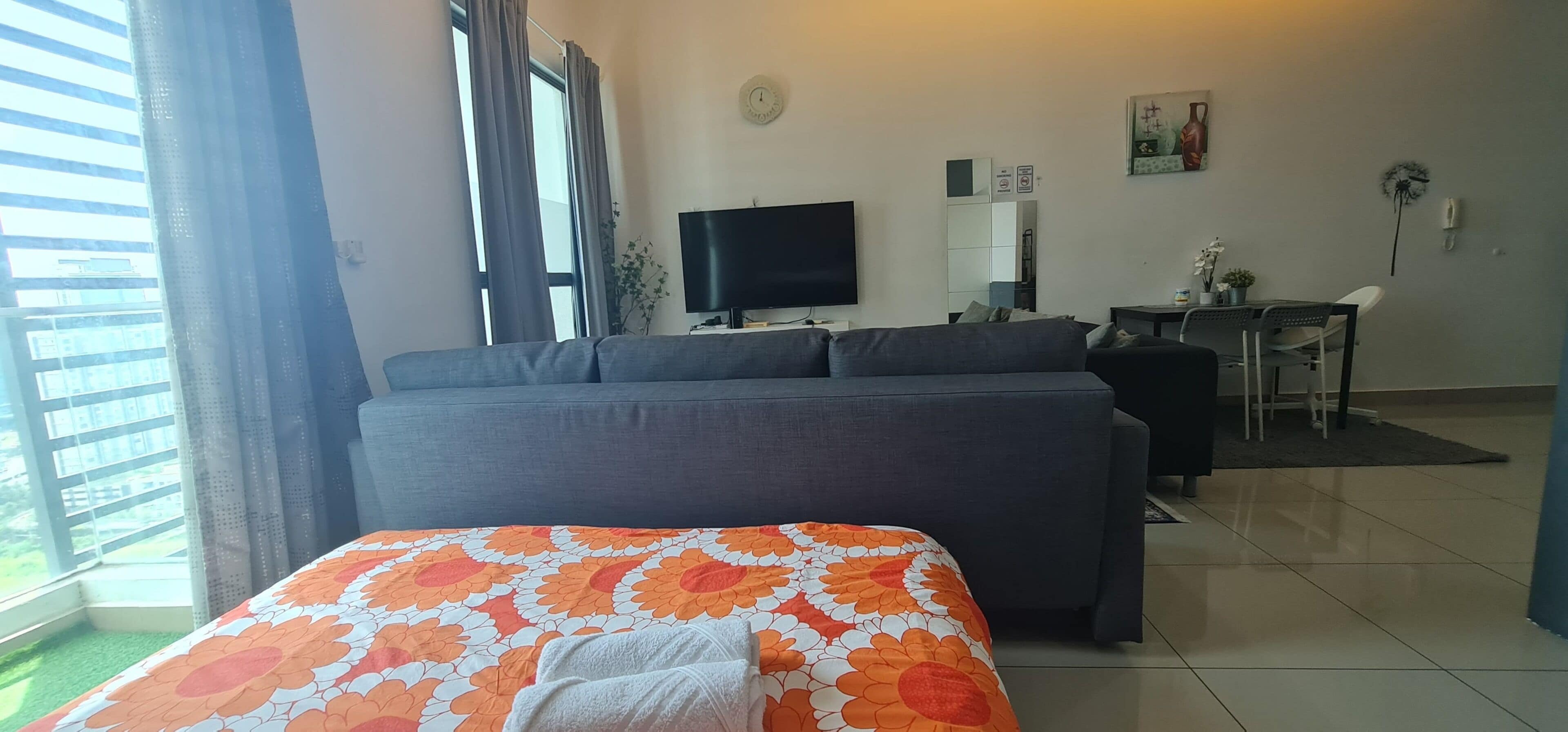 A modern living room with a grey sofa, a flat-screen TV, a dining table with chairs, and a large window with curtains.