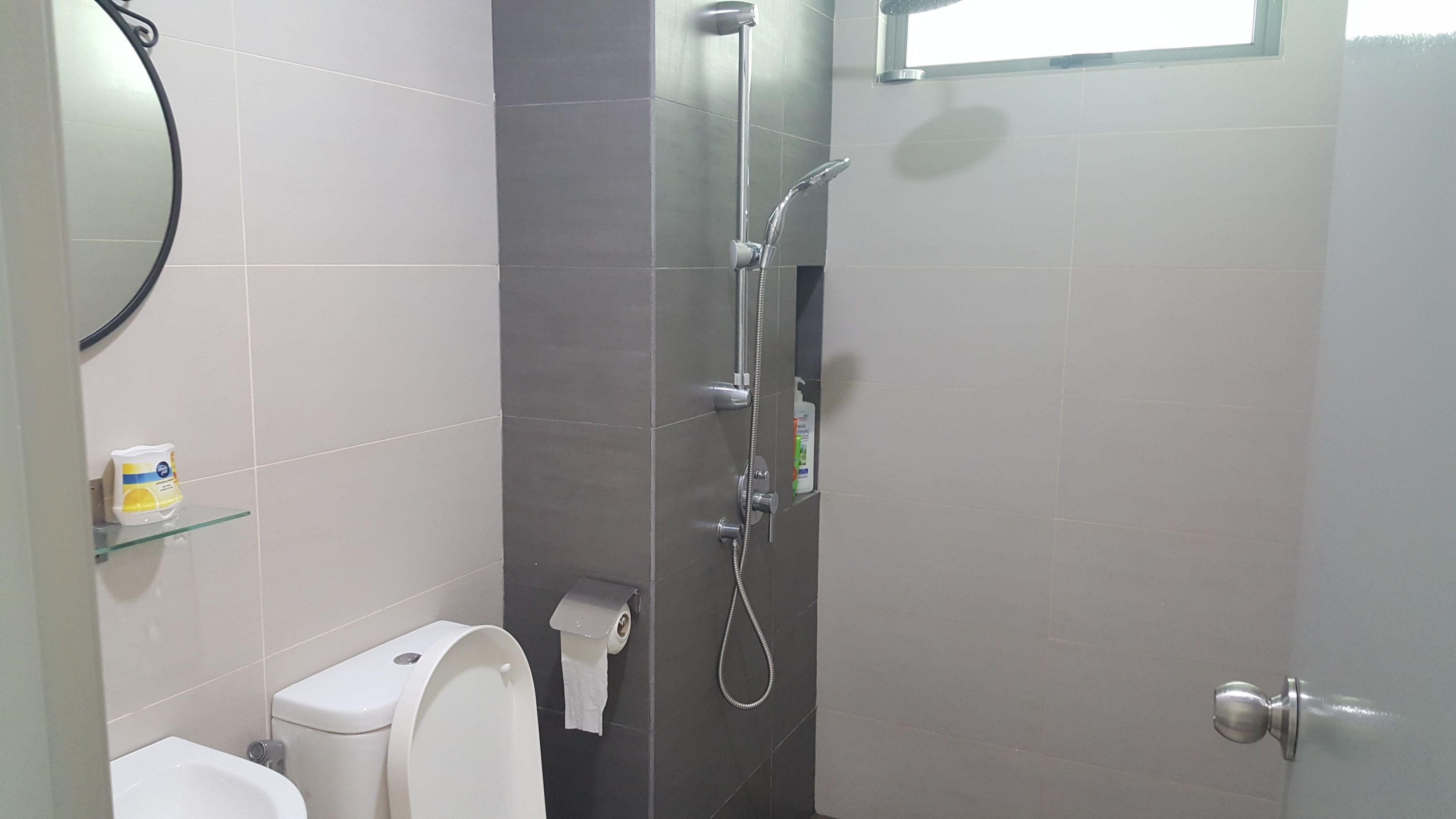 A bathroom with a toilet, a glass shelf with toiletries, a shower with a handheld showerhead, and a towel hanging on the wall.