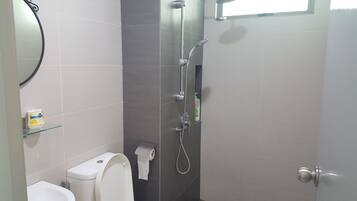 A bathroom with a toilet, a glass shelf with toiletries, a shower with a handheld showerhead, and a towel hanging on the wall.