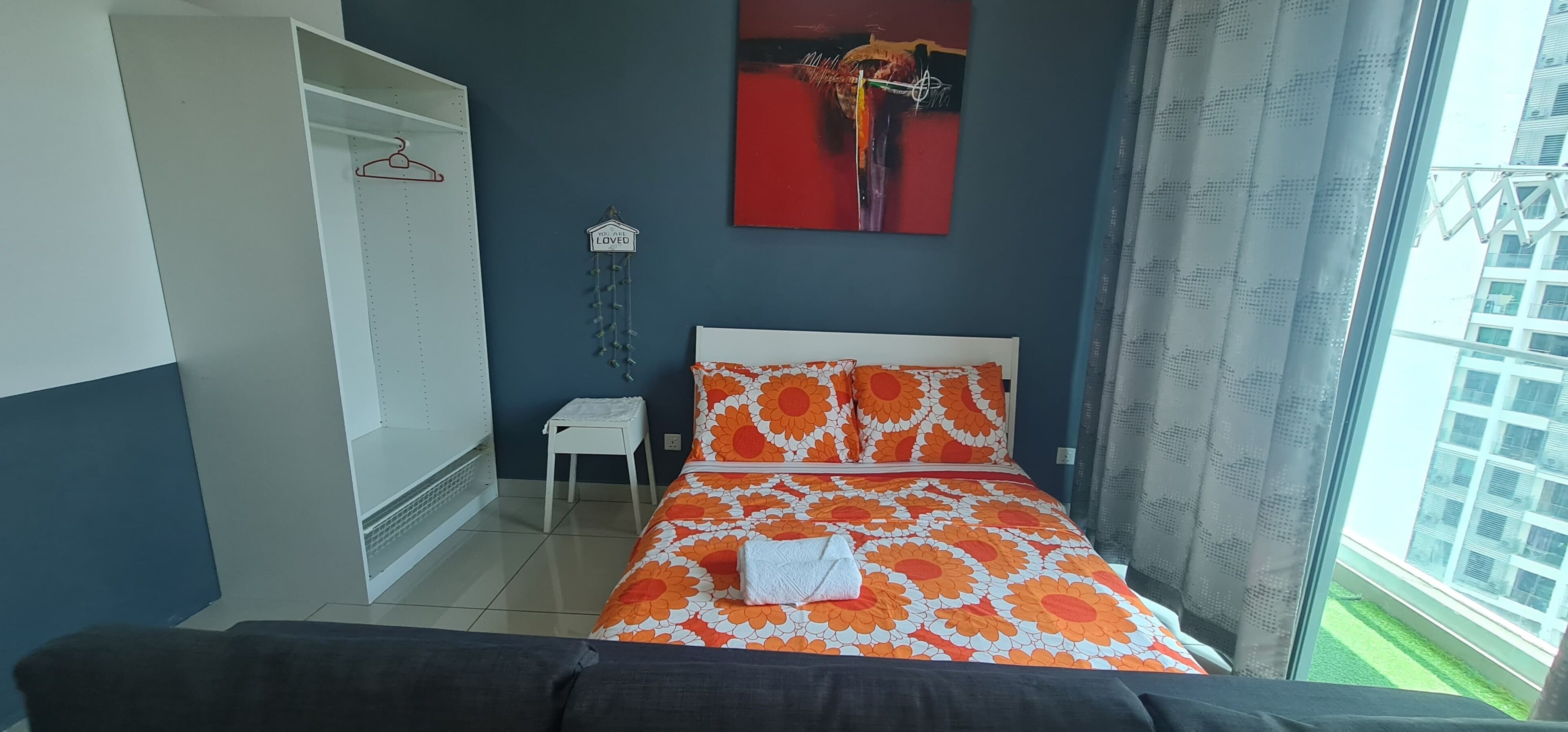 A bedroom with a bed, a wardrobe, a small table, and a window with a view of buildings.