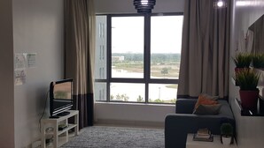 A modern living room with a large window, a flat-screen TV, a sofa, and a small table with books and a plant. - Cyberjaya Vega Residensi (Cyberjaya)