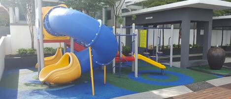 A playground with slides and climbing structures, surrounded by greenery and modern buildings.