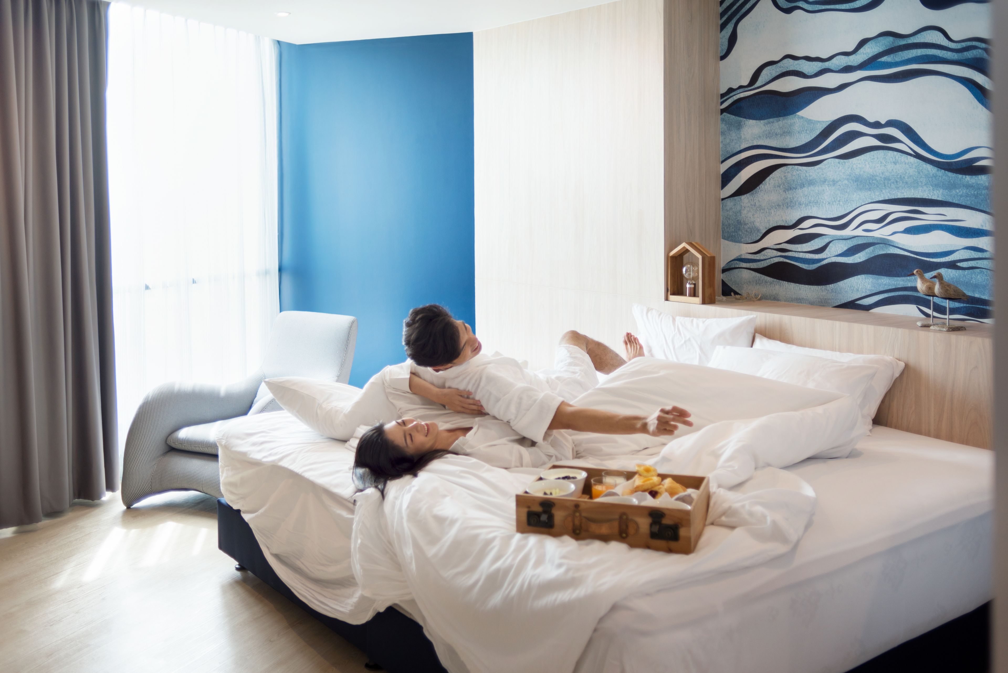 A hotel room with a bed, a bedside table with a lamp, a window with curtains, and a wall with a blue and white abstract painting.