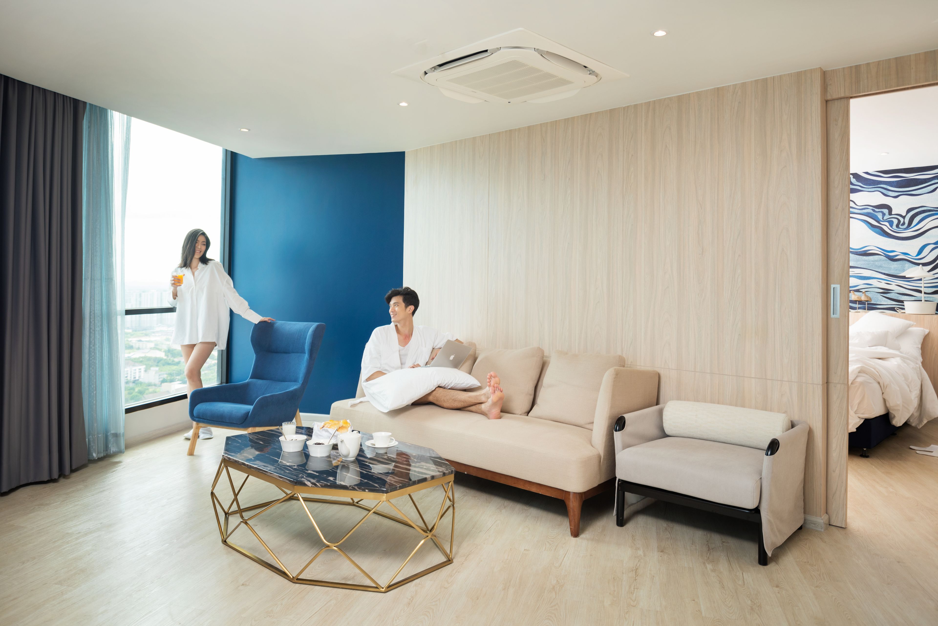 A modern hotel room with a large window, a blue accent wall, a beige sofa, a blue armchair, and a marble coffee table.