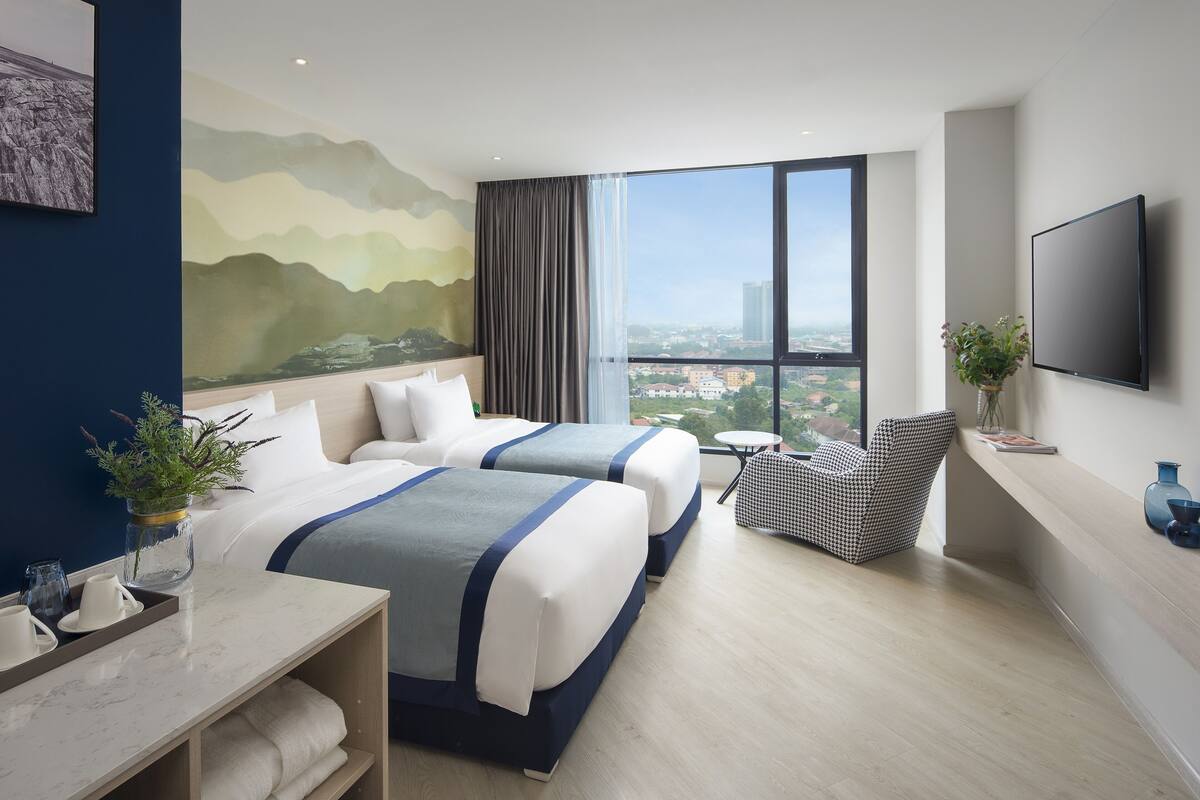 Ein Hotelzimmer mit zwei Betten, einem großen Fenster mit Blick auf die Stadt, einem Flachbildfernseher und einem kleinen Tisch mit Stuhl.