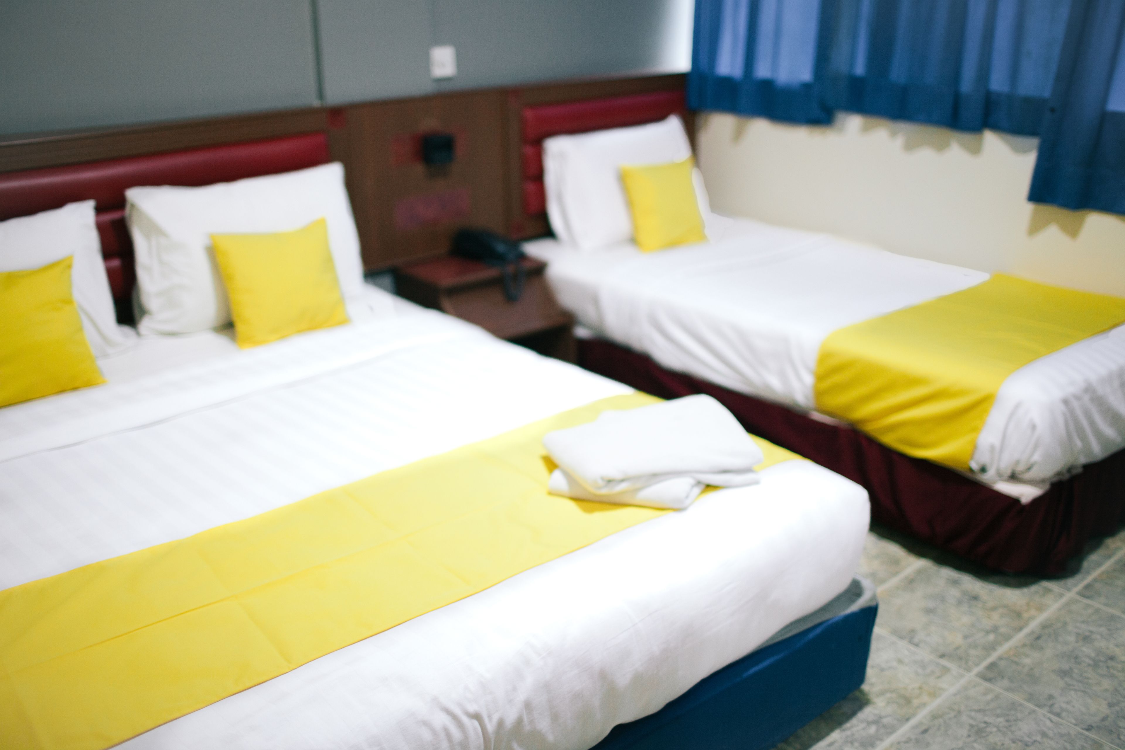 A hotel room with two beds, each with white bedding and yellow accents. There is a blue suitcase on the floor.
