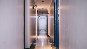 Un pasillo largo y estrecho con suelo de baldosas brillantes, una puerta azul a la derecha y una lámpara a la izquierda.