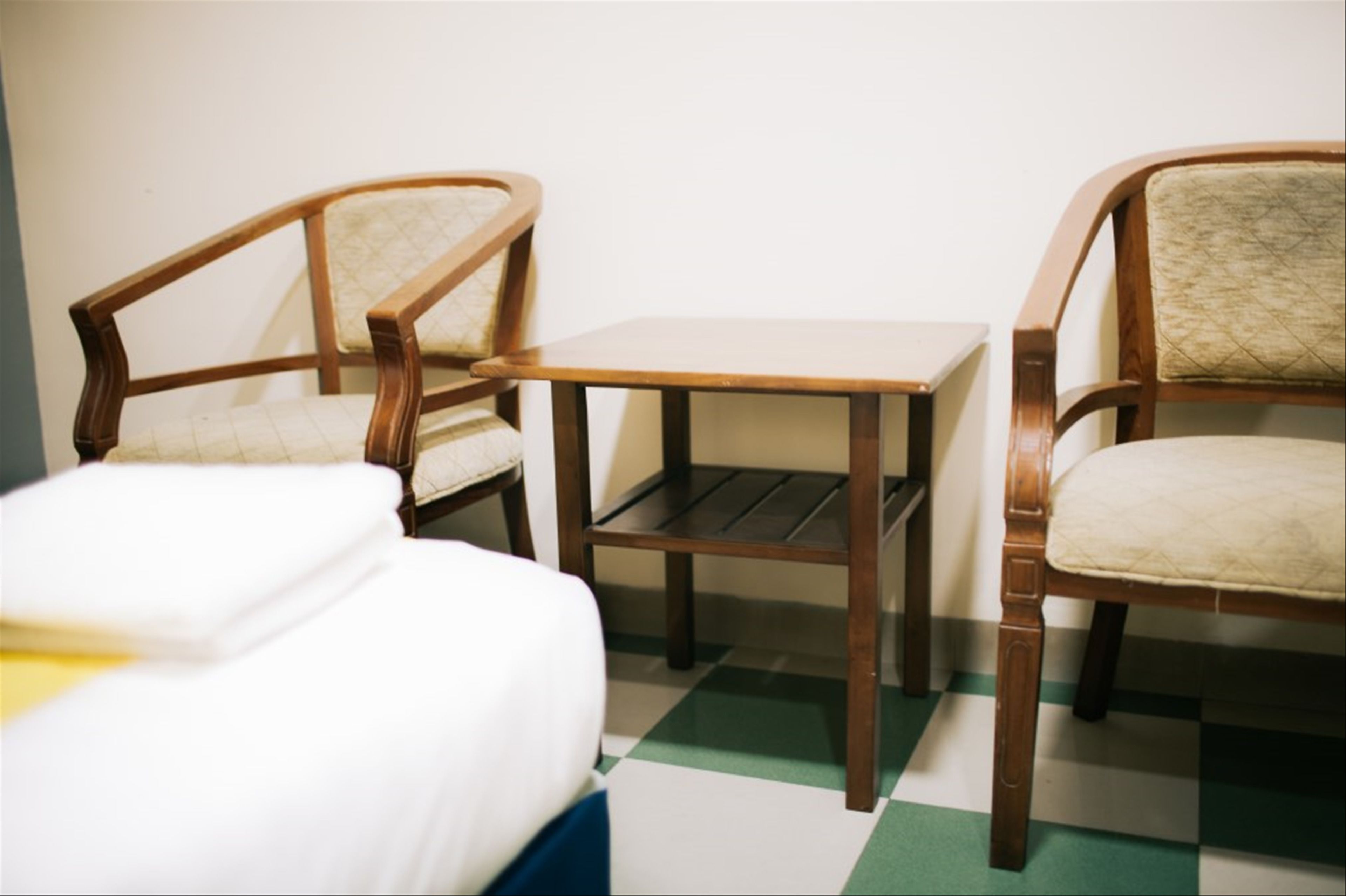 A room with two wooden chairs and a small table, a bed with white bedding, and a checkered floor.