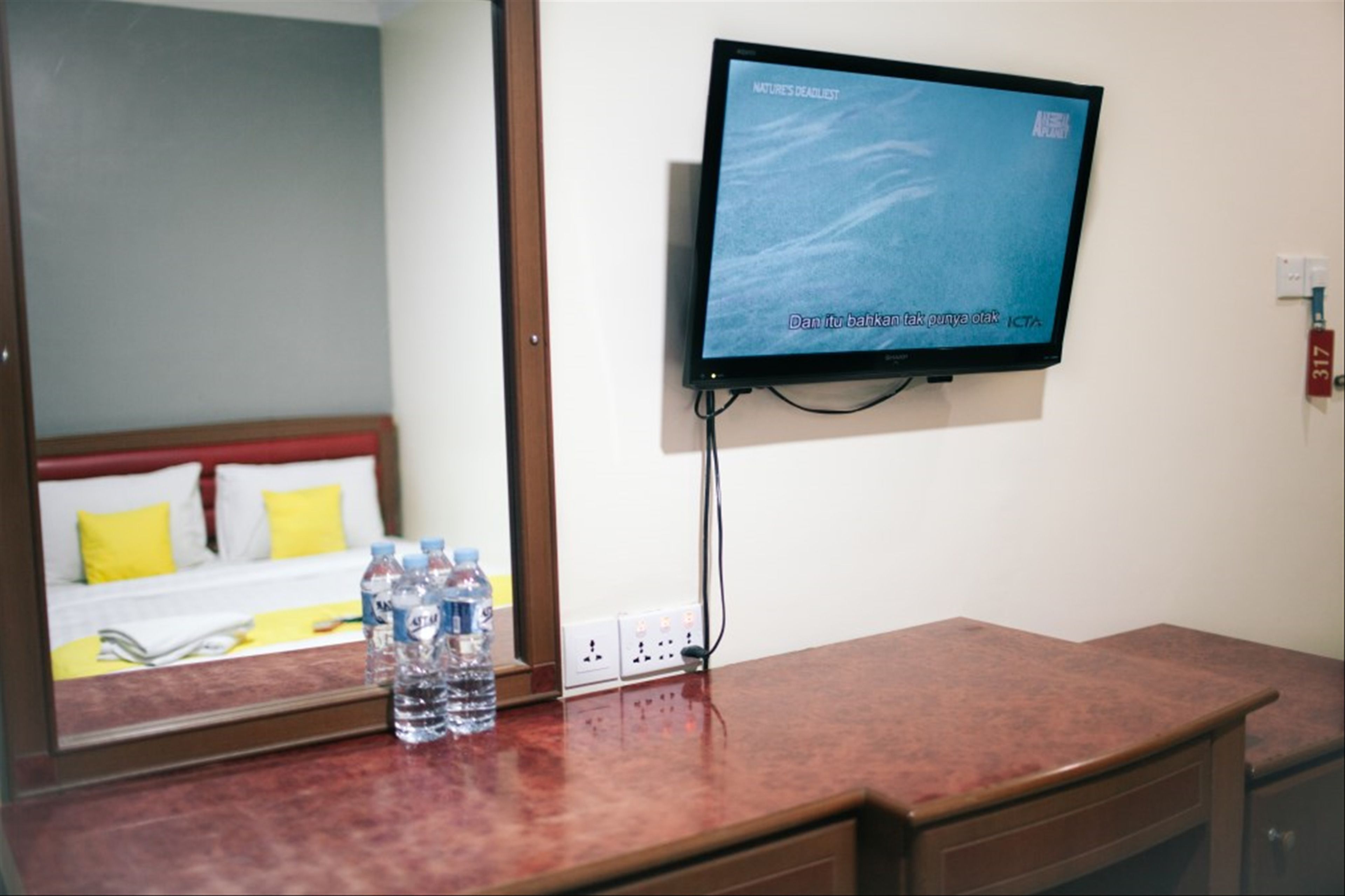 A hotel room with a TV mounted on the wall, a mirror reflecting the room, a wooden desk with a water bottle, and a bed with yellow pillows.