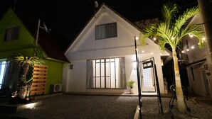 Uma casa de dois andares com telhado inclinado, uma janela grande com cortinas e um pequeno alpendre.