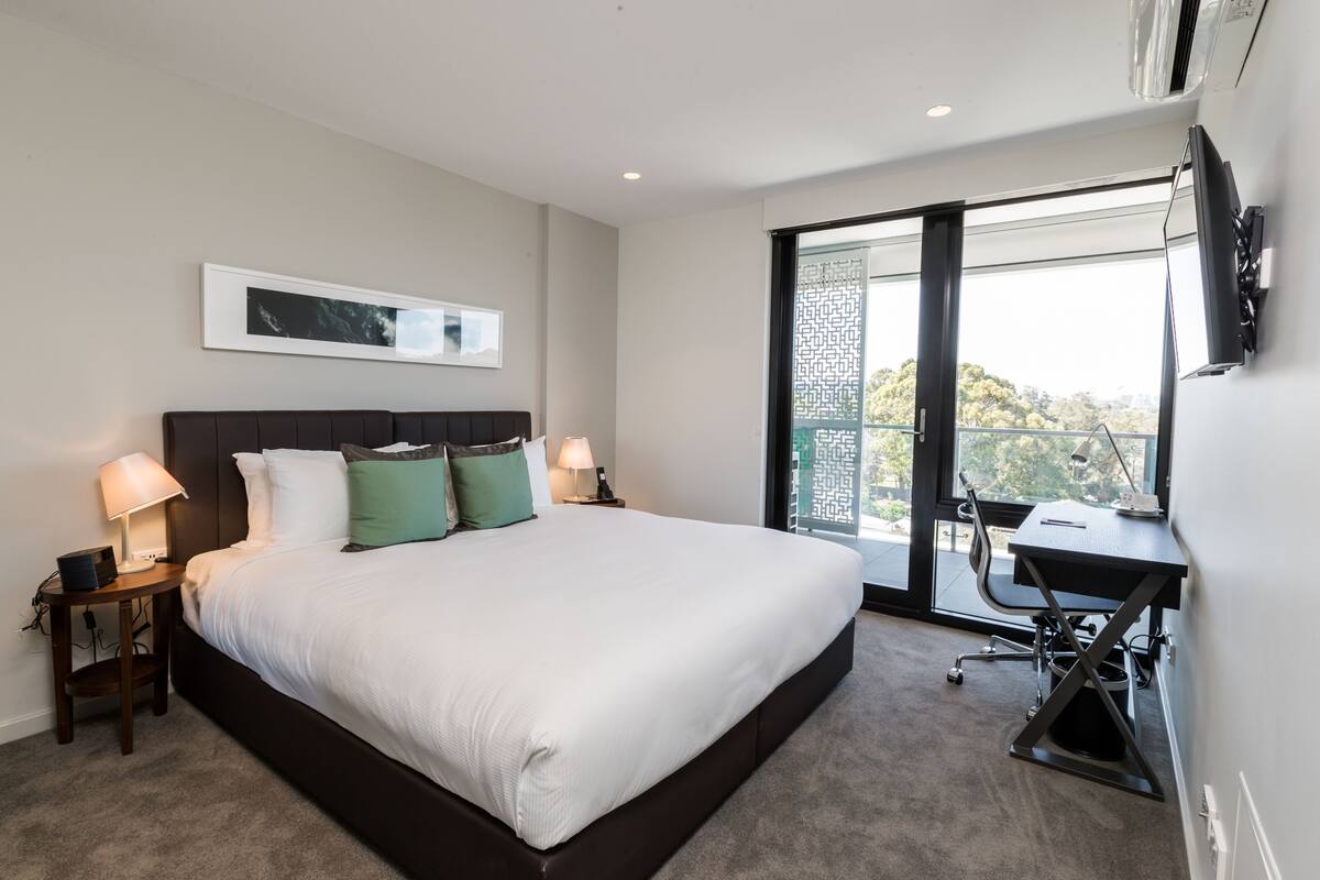 A bedroom with a large bed, a desk, and a view through a sliding door.