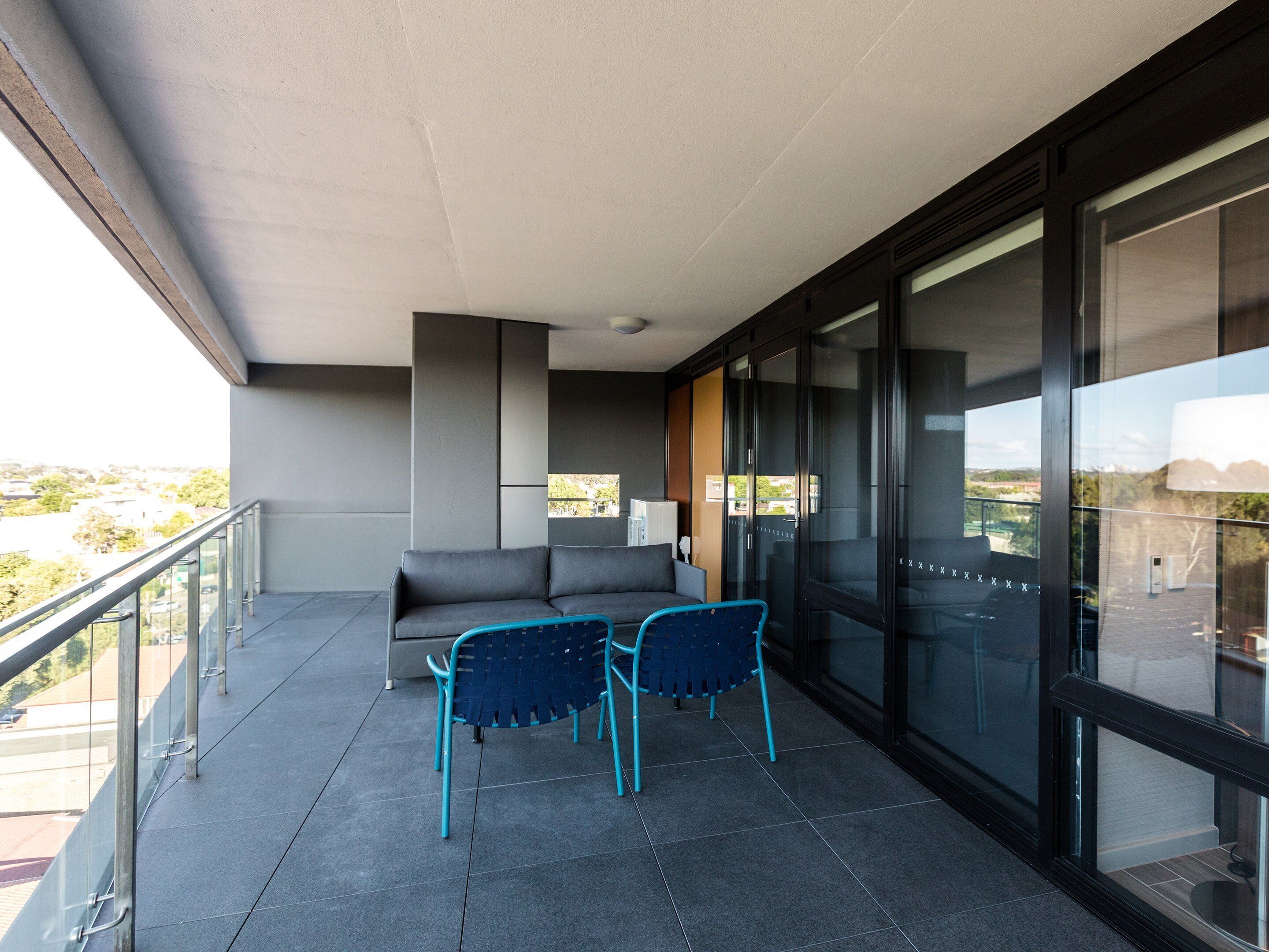 A balcony with a grey sofa and two blue chairs, a glass door, and a view of the city.