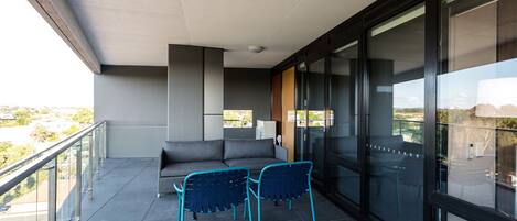 A balcony with a grey sofa and two blue chairs, a glass door, and a view of the city.