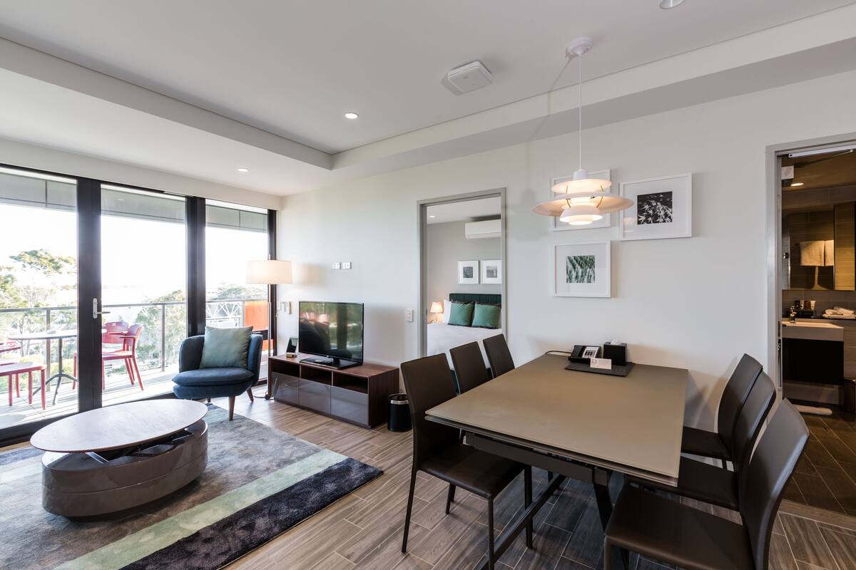 A modern hotel room with a dining area, a TV, and a balcony with red chairs.