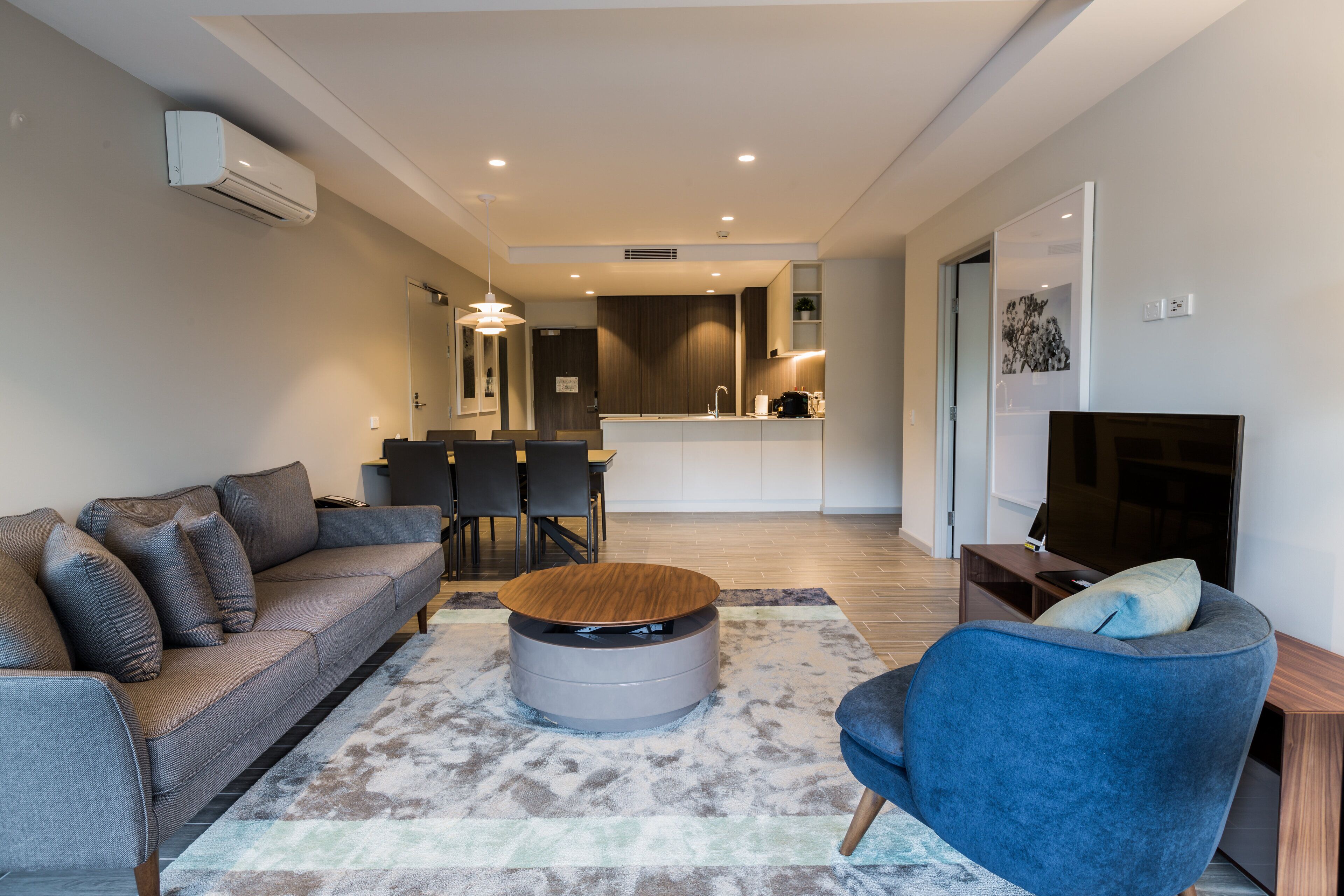 Una sala de estar moderna con un sofá gris, un sillón azul y una mesa de centro redonda. Se observa un comedor con mesa y sillas al fondo.
