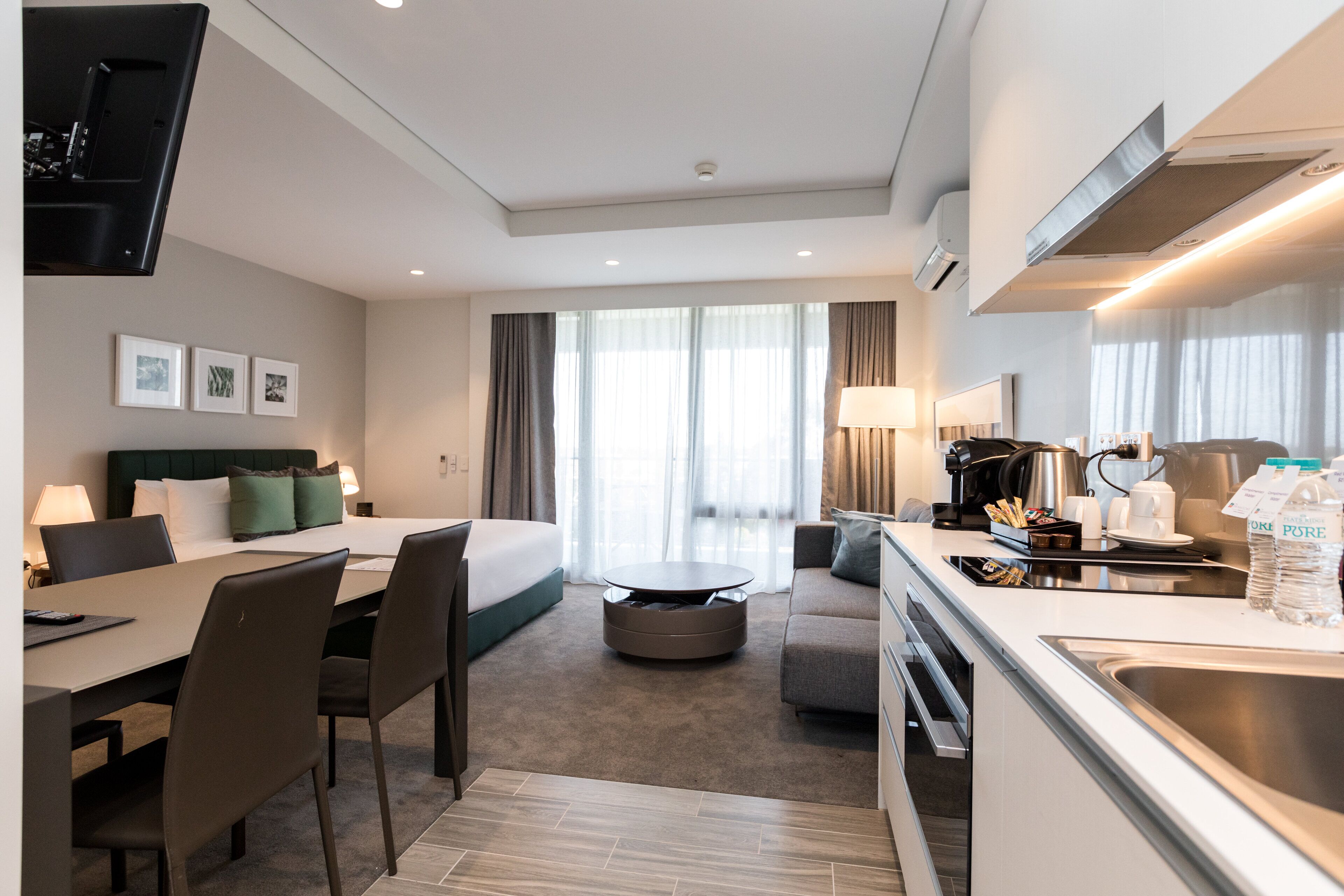 Una habitación de hotel moderna con cama, zona de comedor, cocina y un amplio ventanal con cortinas.