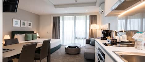 Una habitación de hotel moderna con cama, zona de comedor, cocina y un amplio ventanal con cortinas.