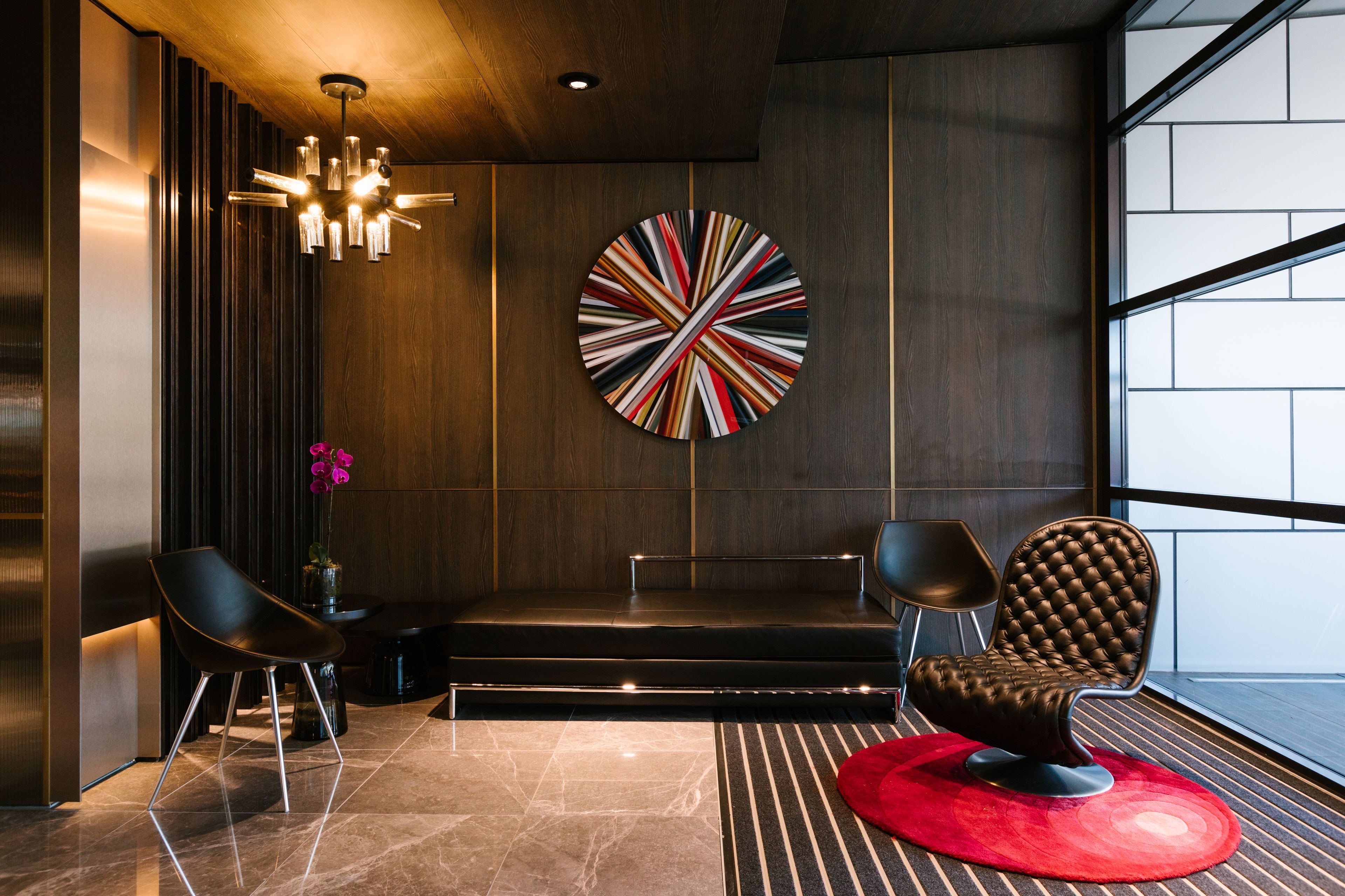 A modern interior with a dark wood panel wall, a round abstract art piece, two black chairs, a red rug, and a ceiling light fixture.