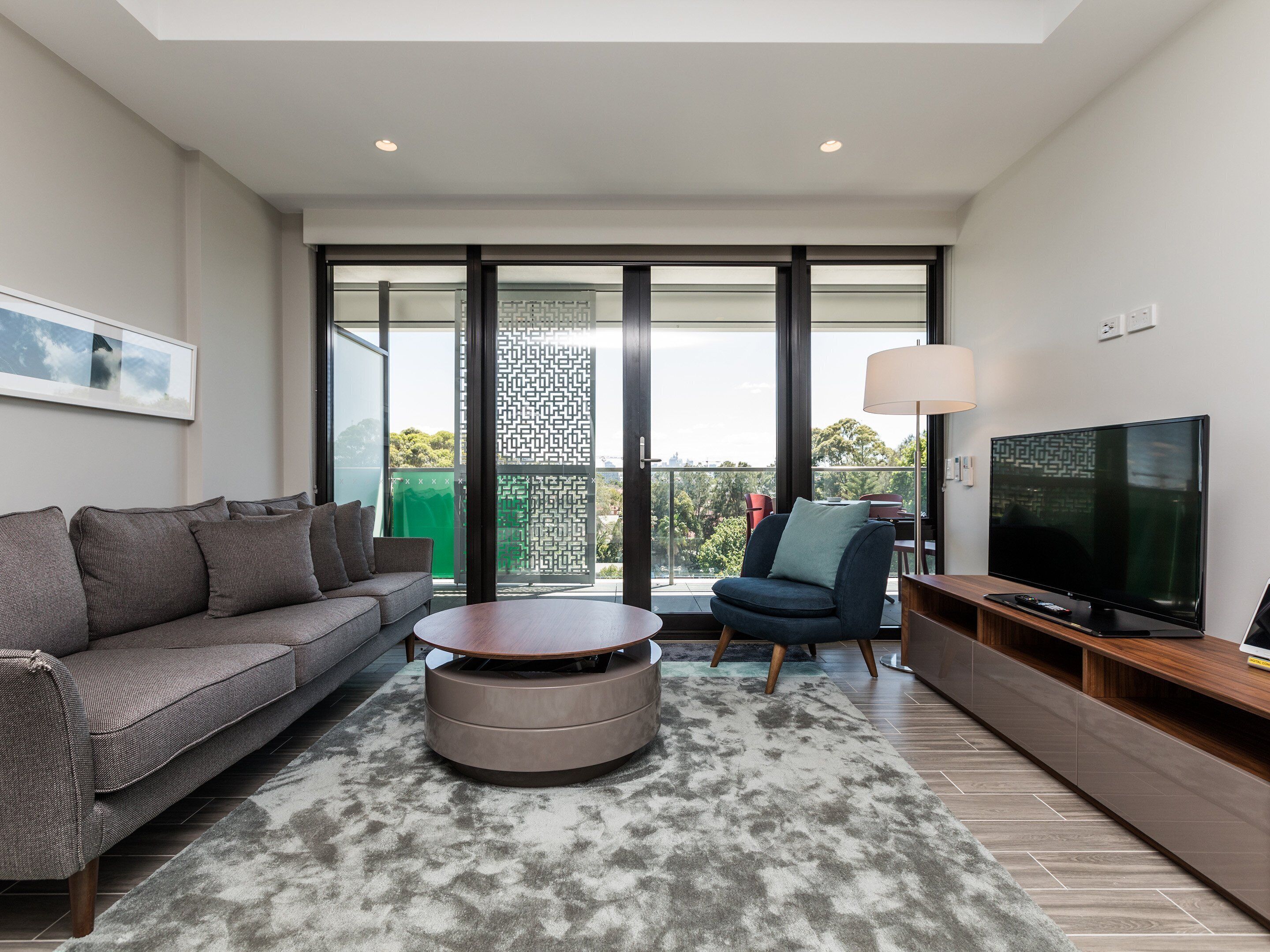 A modern living room with a grey sofa, a round coffee table, a blue armchair, and a TV on a wooden stand.