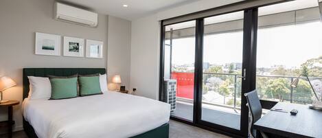 A modern hotel room with a large bed, a desk, and a view of the city through large windows.