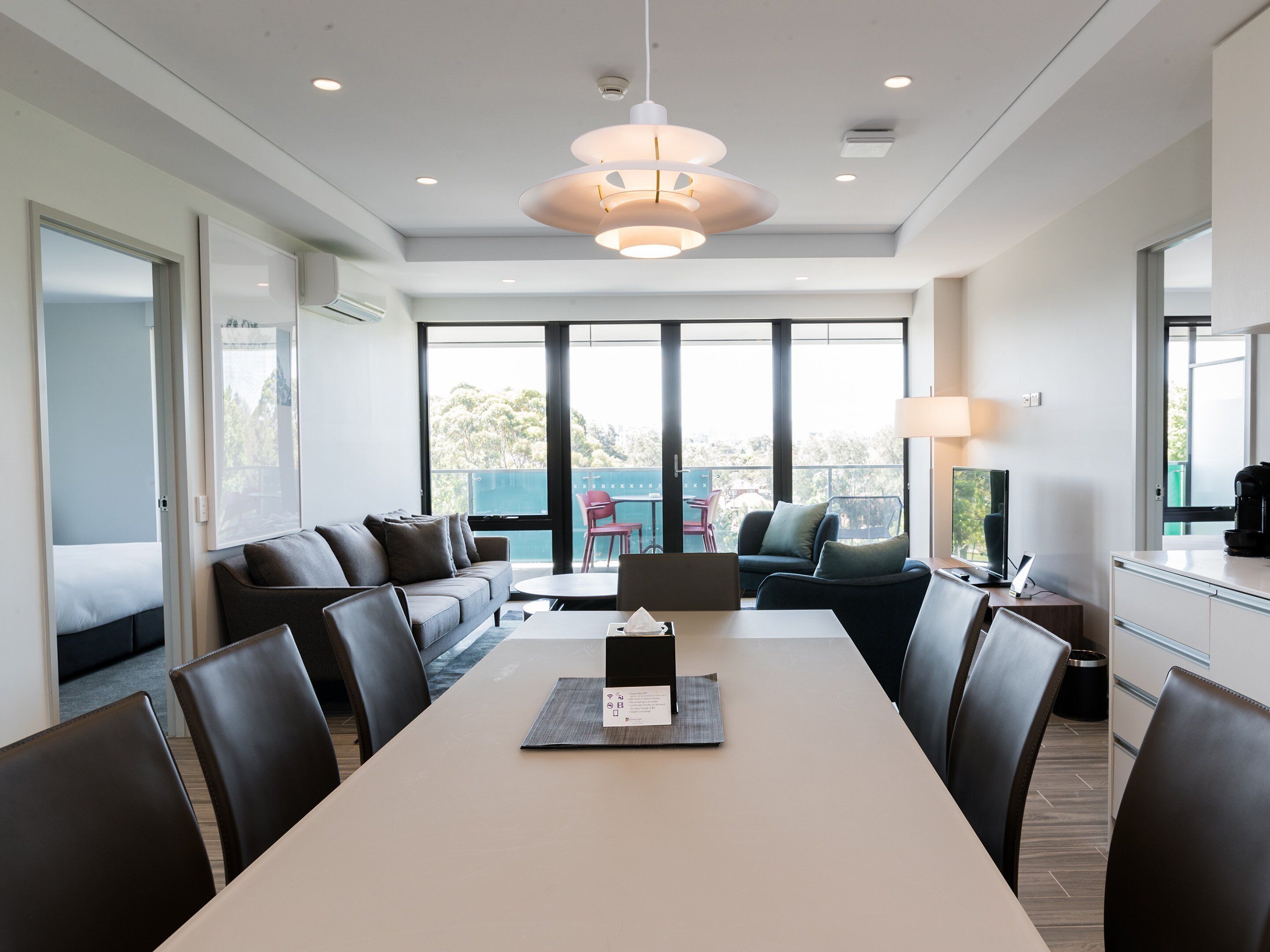 Una habitación de hotel moderna con una amplia mesa de comedor, asientos cómodos, un balcón con vistas y una kitchenette.