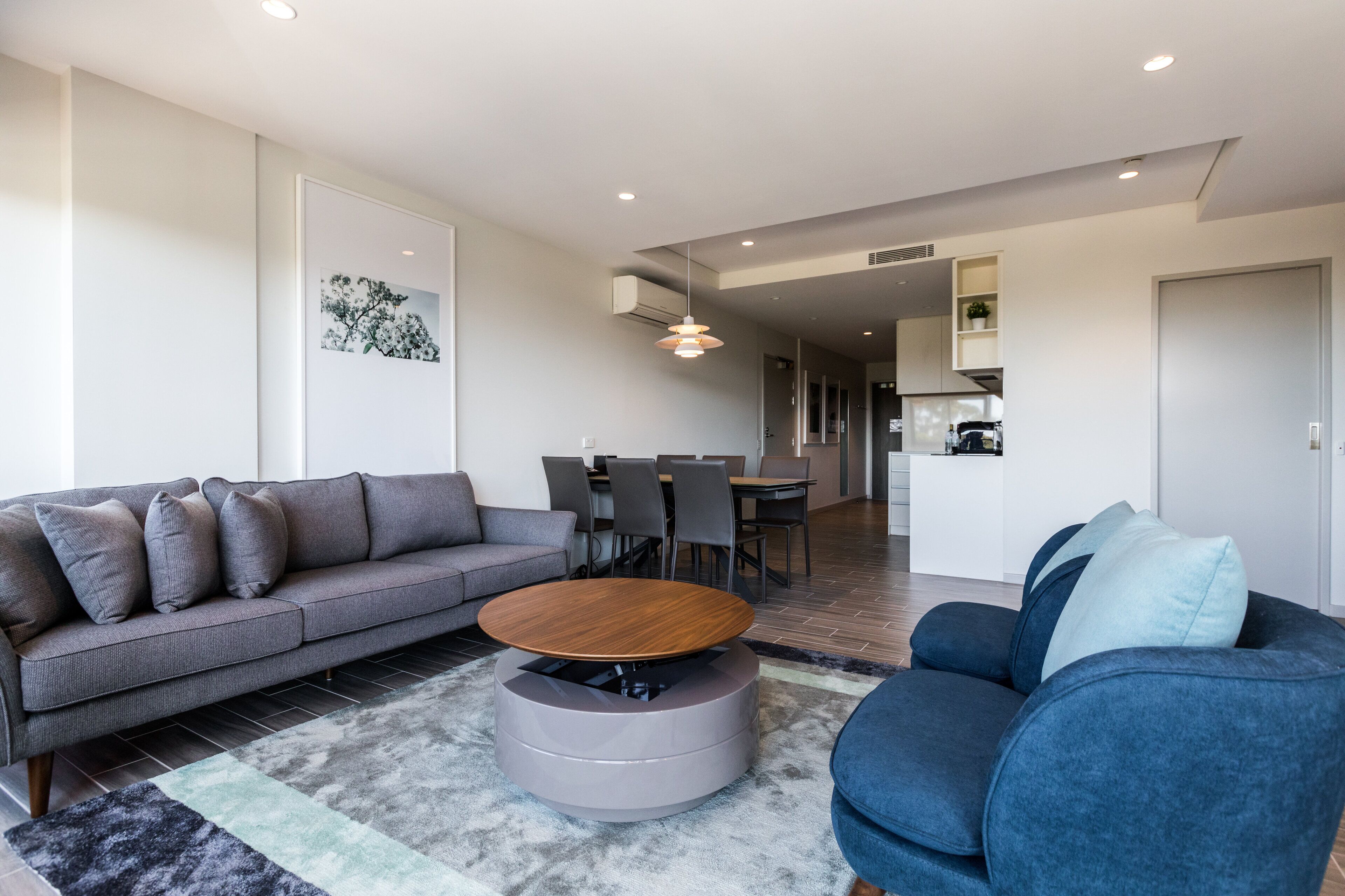 A modern living room with a grey sofa, a blue armchair, a round coffee table, and a dining area in the background.
