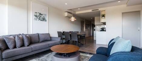 A modern living room with a grey sofa, a blue armchair, a round coffee table, and a dining area in the background.