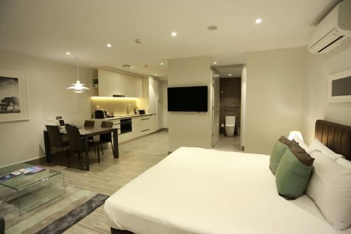 A modern hotel room with a bed, a desk, a chair, a television, and a kitchenette.