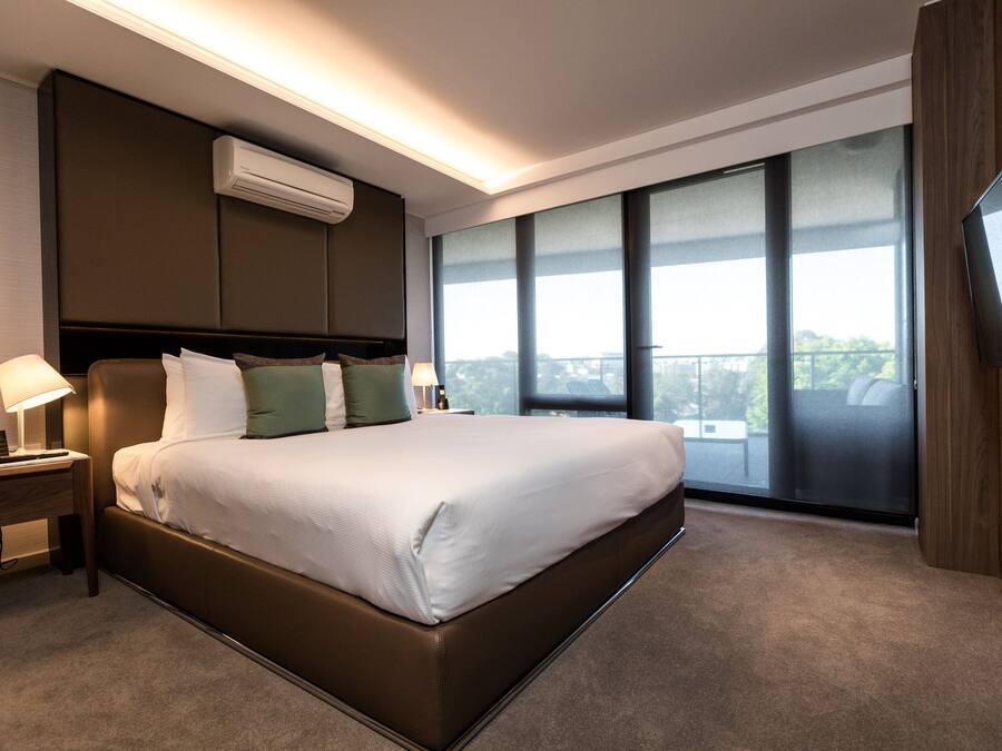 A modern hotel room with a large bed, a nightstand, a lamp, an air conditioning unit, a TV, and a large window offering a view of the outdoors.