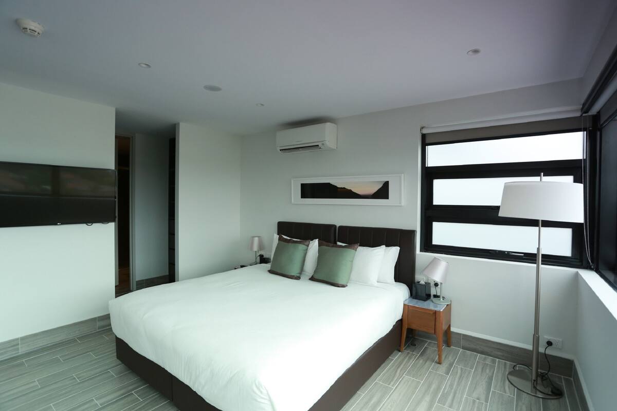 A modern hotel room with a large bed, a nightstand, a floor lamp, and a window with blinds.