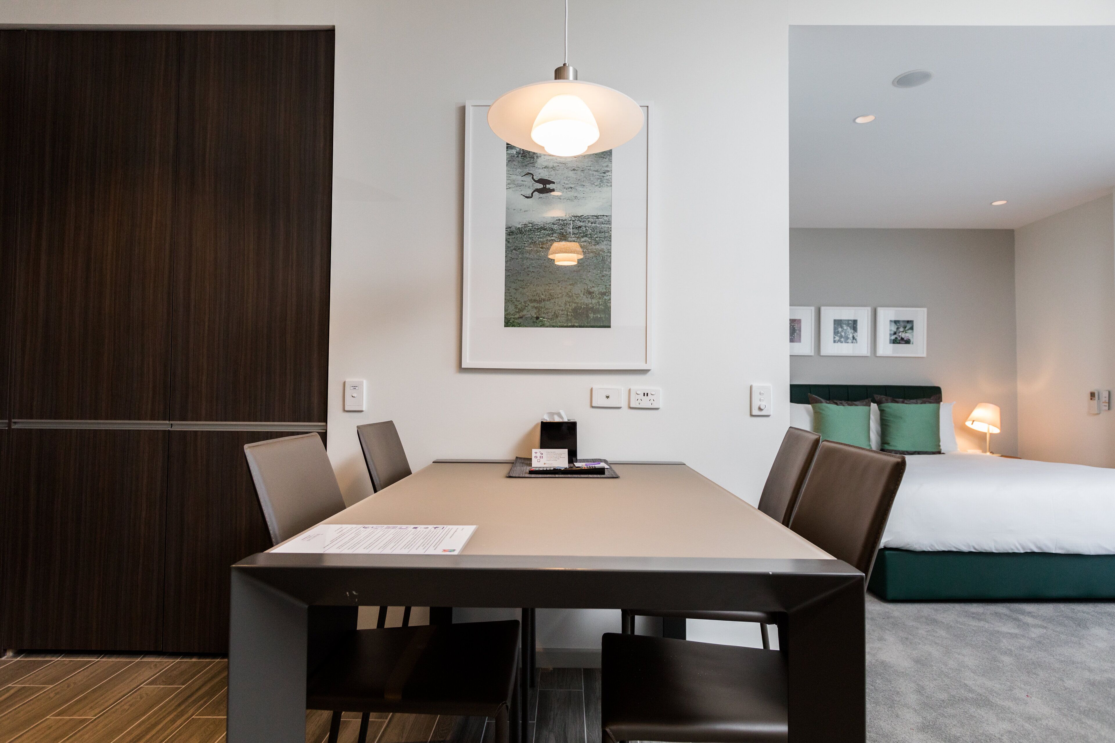 A modern hotel room with a dining area, a bed with green pillows, and a framed picture on the wall.
