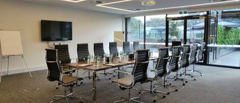 A conference room with a long table, black chairs, a flat-screen TV, and large windows.