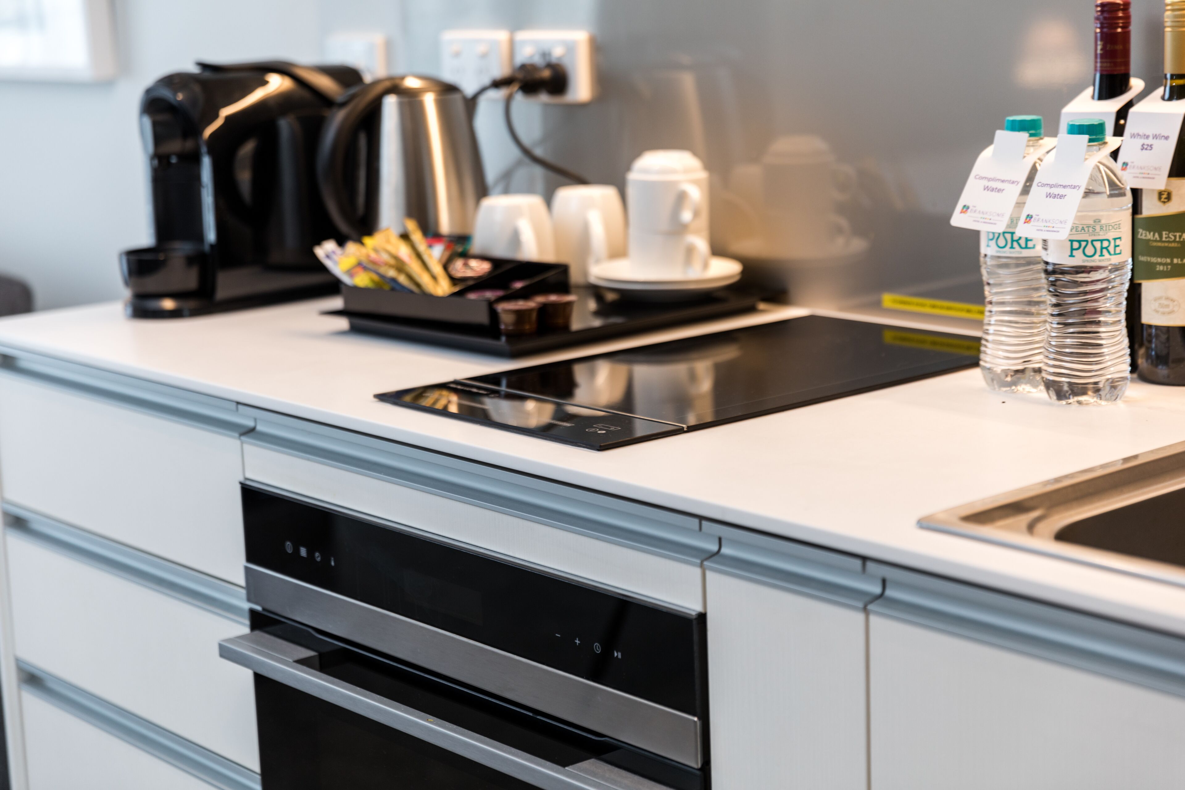A modern kitchen with a built-in coffee maker, a kettle, and a water dispenser. There are also bottles of water and a wine bottle on the counter.