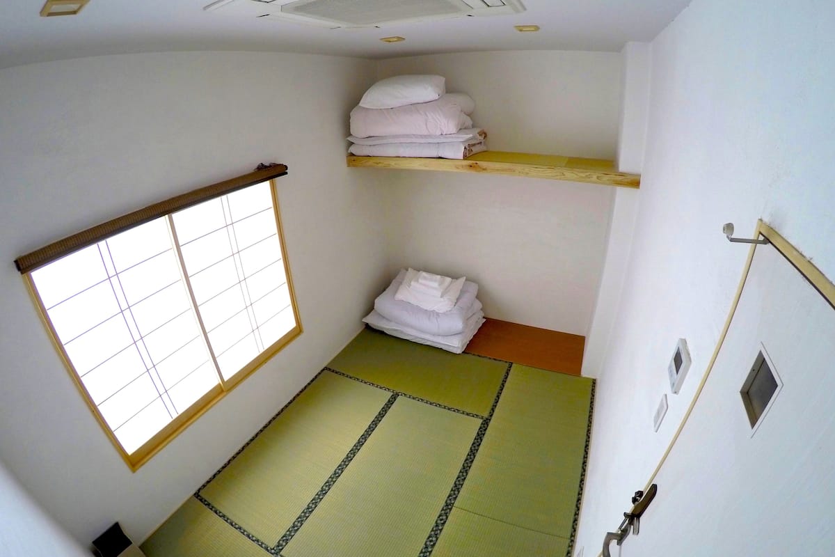 Una habitación con suelo de tatami, un biombos shoji, una estantería con toallas dobladas y una ventana pequeña.