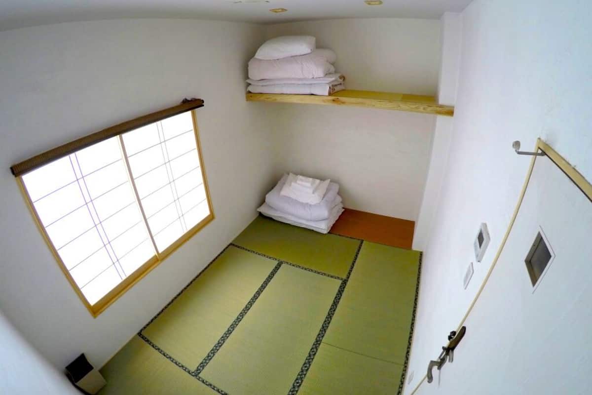 Una habitación con suelo de esteras de tatami, una ventana con diseño de cuadrícula, un estante con toallas dobladas y un pequeño calentador en el suelo.