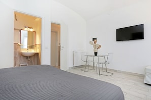 Ein modernes Hotelzimmer mit einem großen Bett, einer kleinen Essecke mit Tisch und Stühlen, einem flachbildfernseher an der Wand und einem Badezimmer mit Waschbecken und Spiegel.